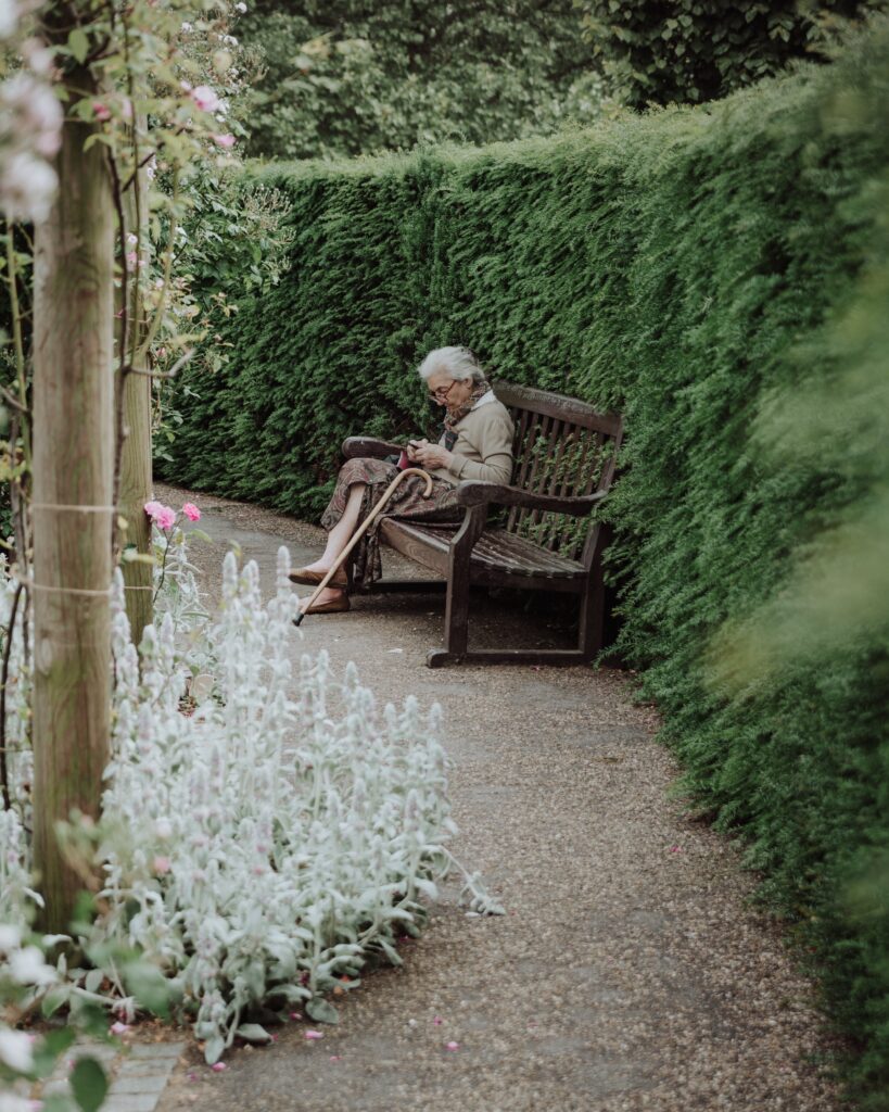 An older woman sitting alone on a bench in a garden with a walking aid next to her